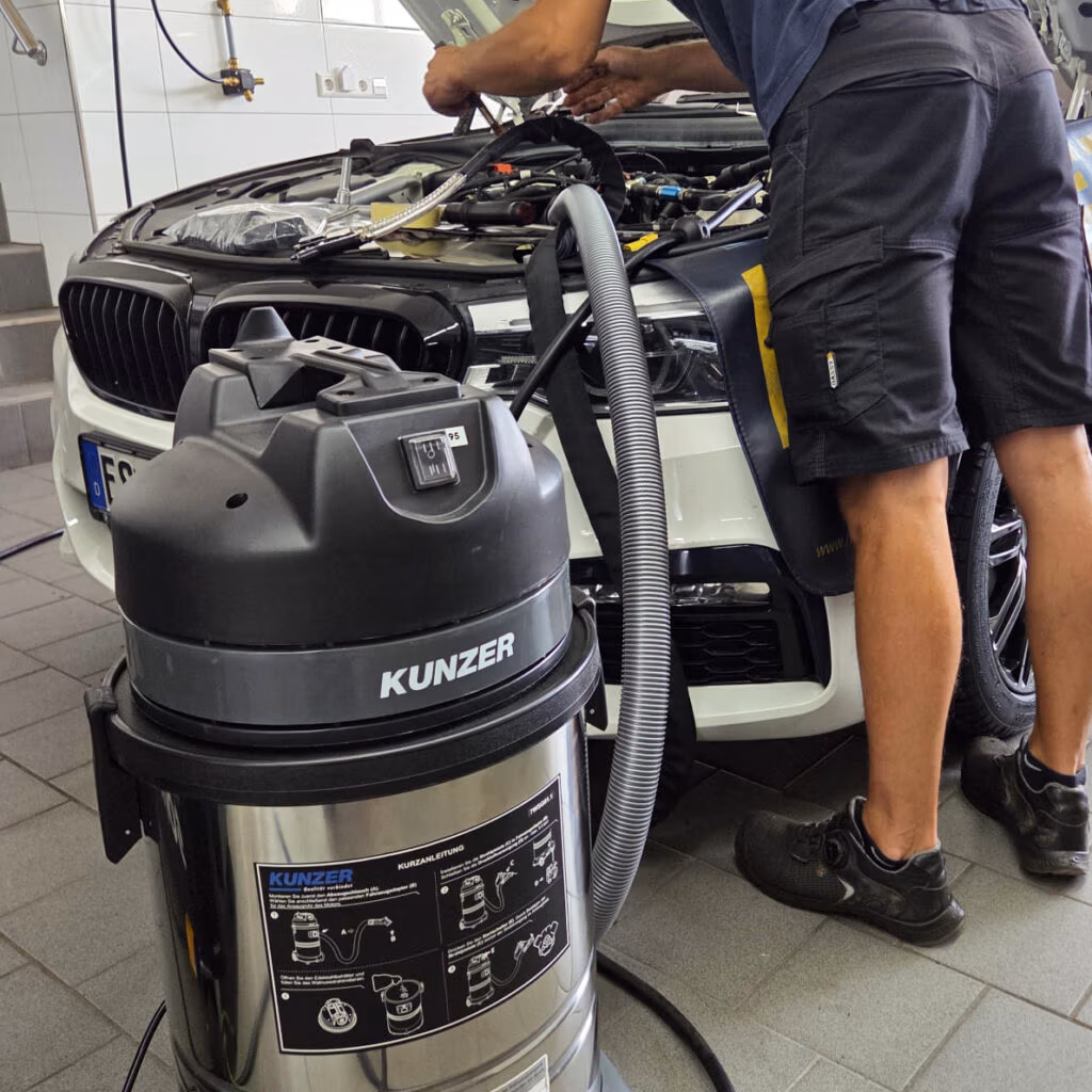 Mechaniker reinigt Einlassventile mit Walnussschalenstrahltechnik; das Kunzer-Strahlgerät steht vor dem Fahrzeug.