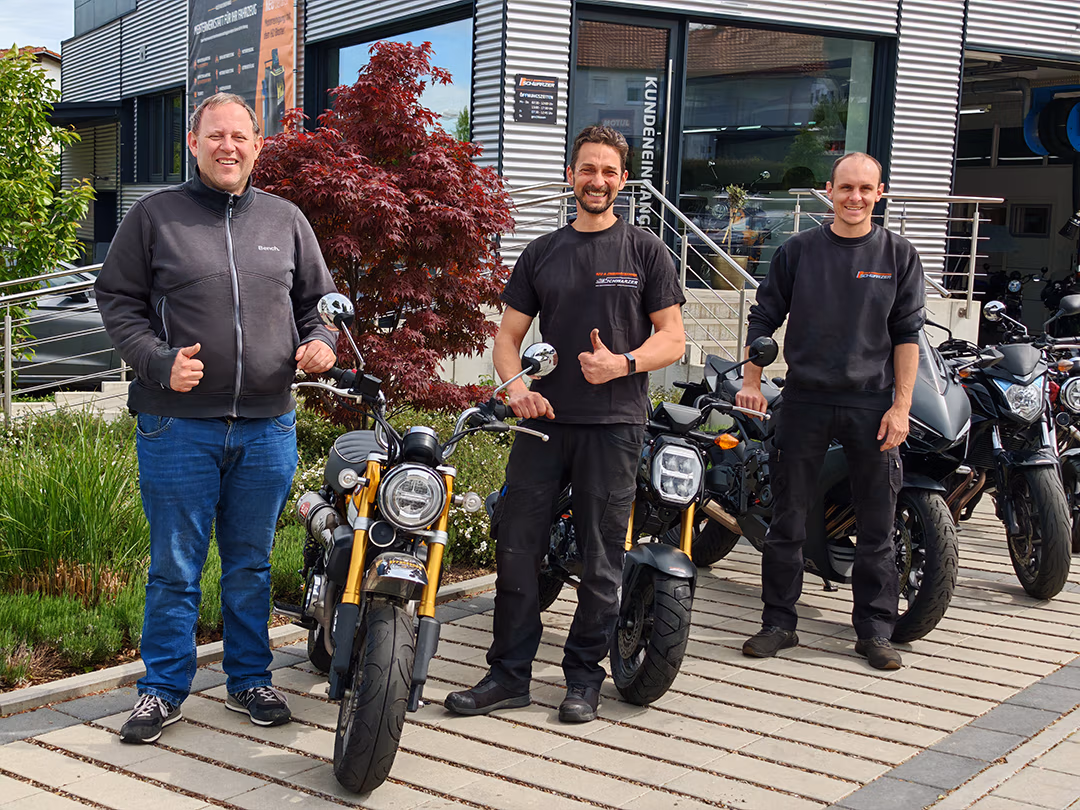 Drei Männer stehen vor der Werkstatt von KFZ Schwarzer neben mehreren Motorrädern. Im Vordergrund sind Honda-Motorräder zu sehen, die im Rahmen der Zusammenarbeit mit Heikos Fahrschulen genutzt werden.